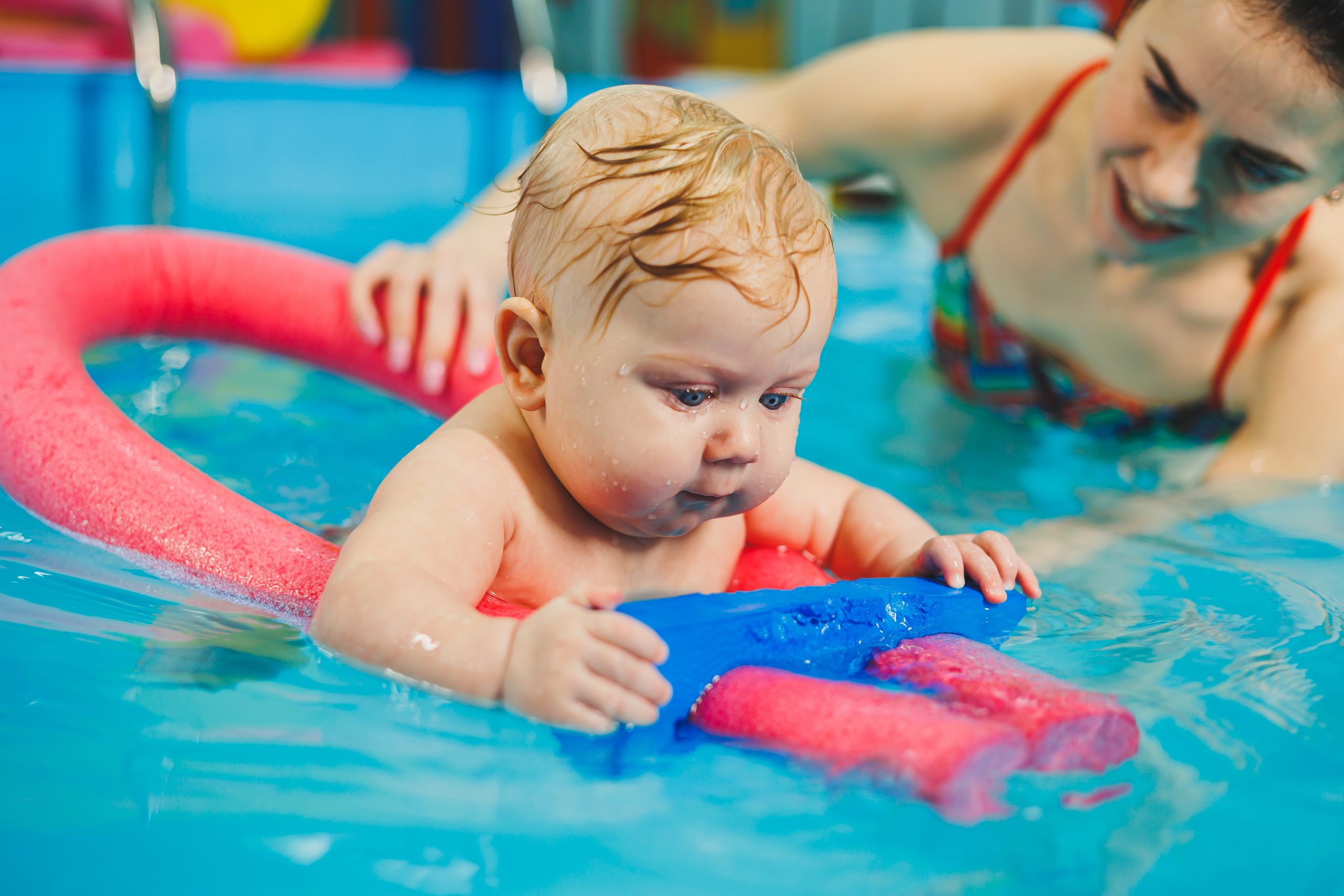 Babyschwimmen in Hamburg: Baby übt Wassergewöhnung mit roter Poolnudel im Schwimmbecken, unterstützt von einer Trainerin
