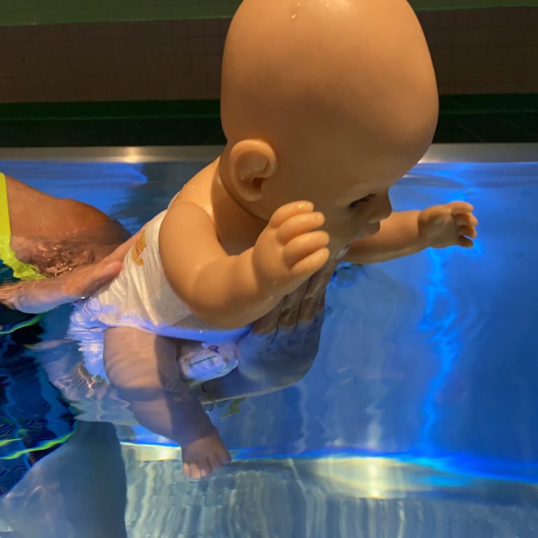 Trainer demonstriert beim Babyschwimmen Hamburg mit einer Puppe die richtige Halteposition im Wasser