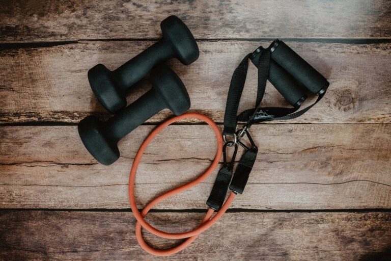 Zwei schwarze Hanteln und ein rotes Fitnessband mit Griffen liegen auf einem Holzboden – Trainingszubehör für Sport und Fitness