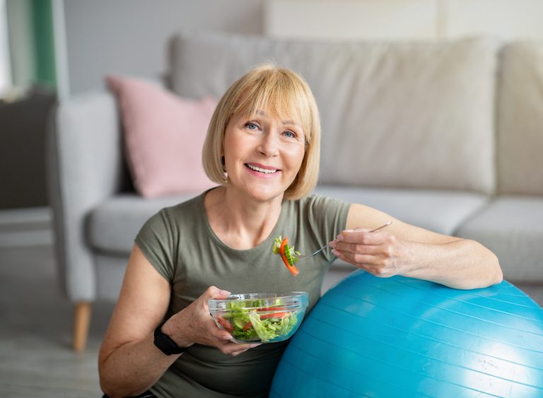 Gesunde ältere Frau sitzt neben Gymnastikball und isst frischen Salat nach dem Training