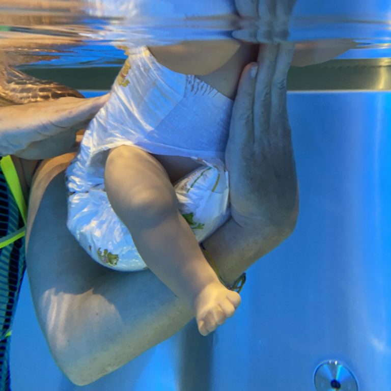 Trainer zeigt beim Babyschwimmen Hamburg mit einer Puppe, wie Babys sicher unter Wasser gehalten werden